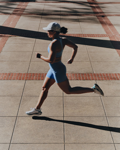 Woman in athletic wear running outside on a brick and concrete surface in bright sunlight, showcasing fitness and an active lifestyle.