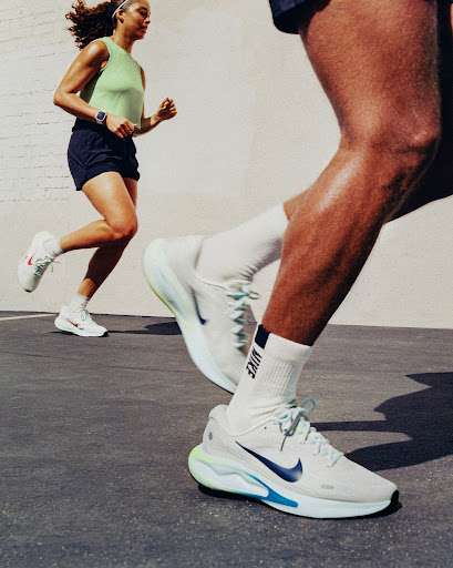 Two athletes running outdoors on pavement; a female runner in the background and a male runner with running shoes in the foreground.