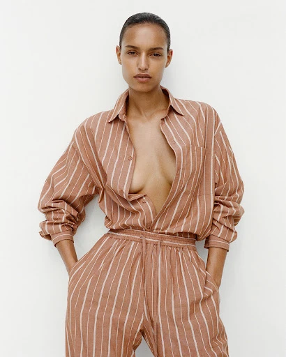 Striking studio shot of a dark-haired adult woman wearing a striped beige two-piece outfit, standing indoors against a plain white wall.