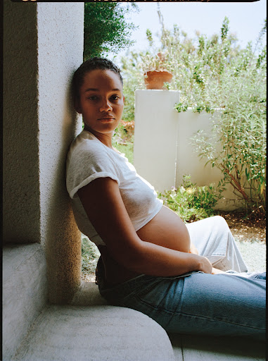 A pregnant woman in a white t-shirt and jeans sits by a window with an outdoor garden background, creating a serene portrait.