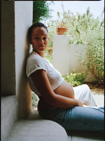 A pregnant woman in a white t-shirt and jeans sits by a window with an outdoor garden background, creating a serene portrait.