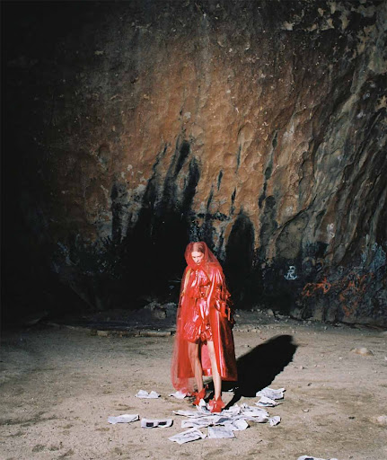 A woman in a red hooded cloak stands among scattered papers in a cave, with a strong light source creating shadows on the cave walls.