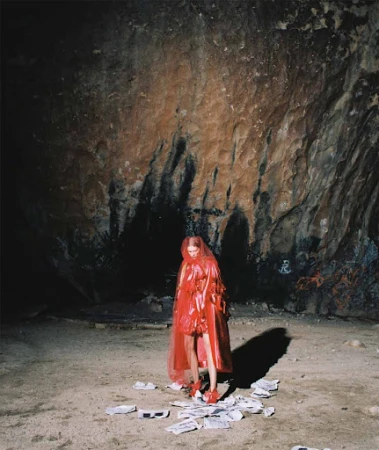 A woman in a red hooded cloak stands among scattered papers in a cave, with a strong light source creating shadows on the cave walls.
