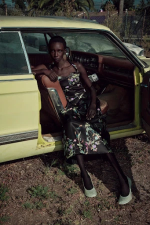 Black woman wearing a floral dress seated inside a vintage yellow car with brown interior.