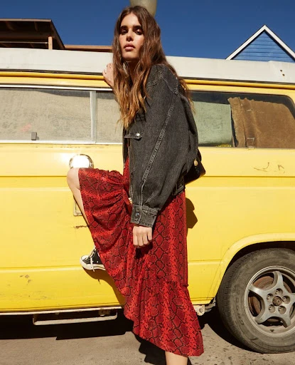 Woman in snakeskin print skirt and denim jacket posing by a yellow van, showcasing fashion and outdoor style in natural light.