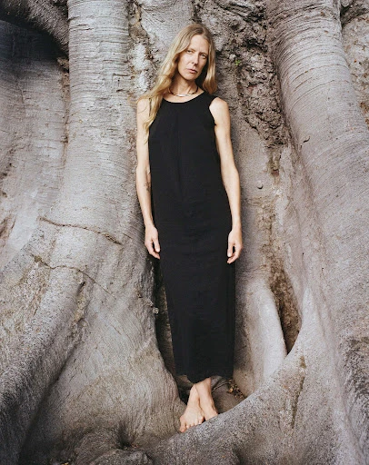 Blonde woman in black dress stands barefoot in the hollow of a large, light gray tree in an outdoor nature setting.