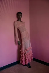 Model wearing a long pink textured knit dress leaning against a dark pink corner wall with soft shadow details.