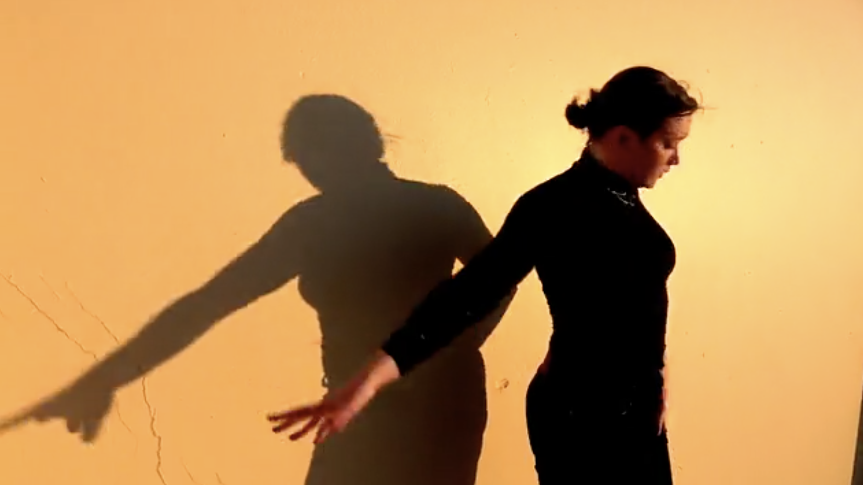 Woman in black clothing stands against a yellow wall, casting a large shadow that mimics her pose in a starkly lit indoor space.