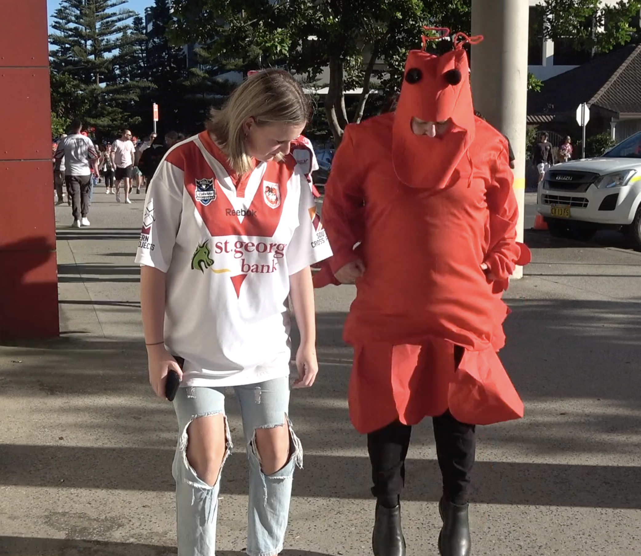 Two people pose on a city street on a sunny day, one wearing an Australian football jersey and the other dressed in a lobster costume.
