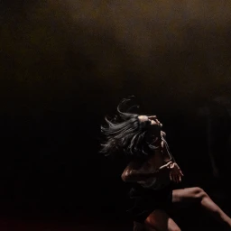 A dancer with dark hair whipped in motion performs under dramatic overhead lighting against a dark low-key background.