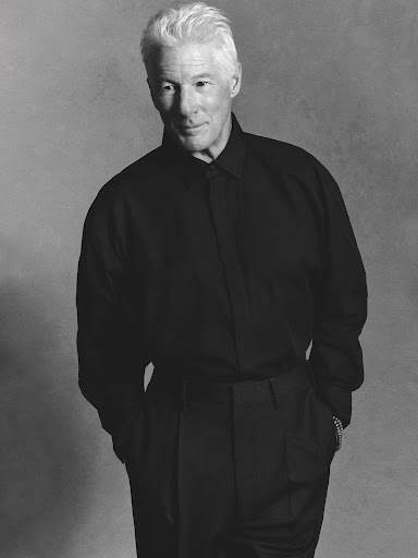 Monochrome portrait of an adult male with short, white hair wearing a dark long-sleeve dress shirt and trousers against a grey backdrop.