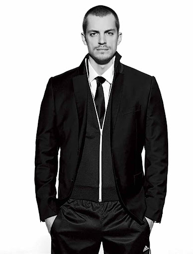 Portrait of an adult male in a black suit jacket, vest with zipper, tie, and black athletic pants against a white background in a studio.
