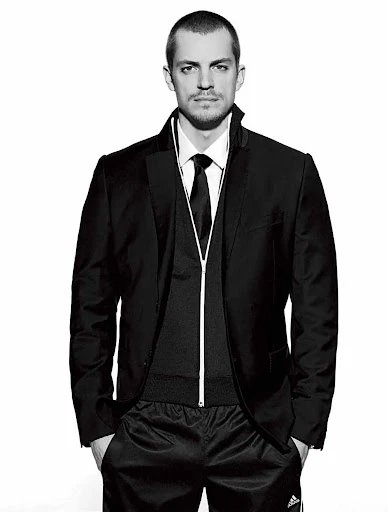 Portrait of an adult male in a black suit jacket, vest with zipper, tie, and black athletic pants against a white background in a studio.