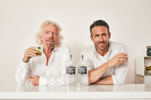 Richard Branson and Ryan Reynolds pose indoors with Aviation Gin bottles on a white counter with bar accessories.