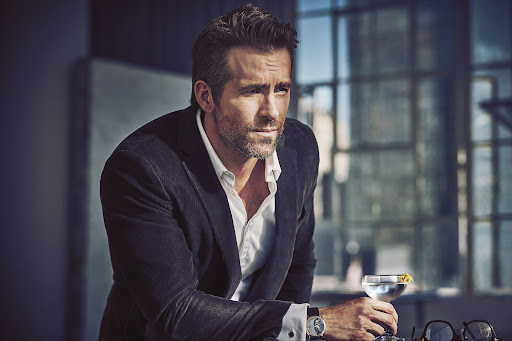 Actor Ryan Reynolds wearing a dark blazer and white shirt holding a cocktail glass indoors