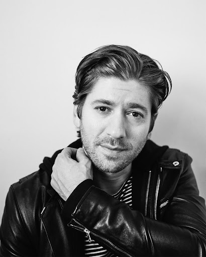 Black and white portrait of an adult male in a leather jacket with a hand near his neck, showcasing a classic yet modern aesthetic.