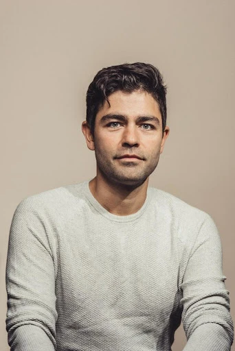 Portrait of a brunette adult male with a neutral expression, wearing a gray textured sweater against a beige background. Head and shoulders shot.