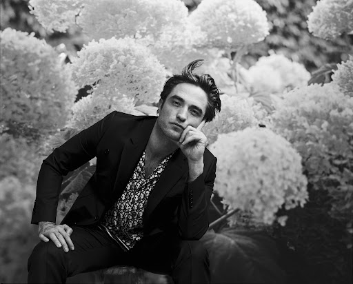 Portrait of Robert Pattinson in a black suit and patterned shirt sitting in front of large white flowers looking at the camera.