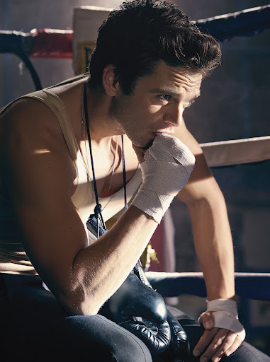 A man with wrapped hands sits in a boxing ring corner, wearing a tank top and boxing gloves, with a thoughtful expression. 