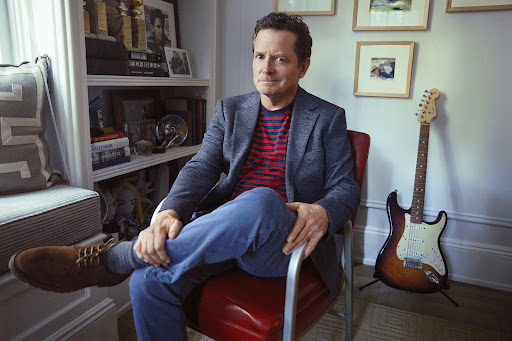 Portrait of Michael J. Fox sitting in a red chair, wearing a blazer, striped shirt, jeans, and brown shoes in a home setting.
