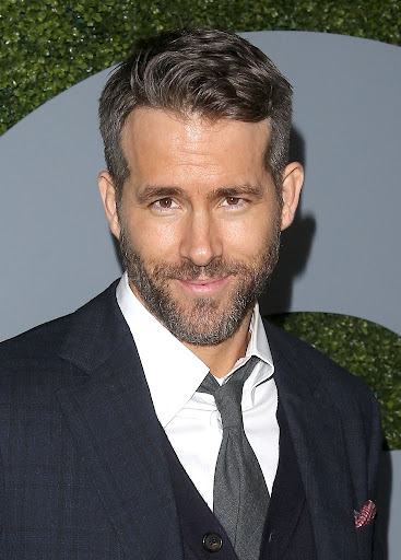 Ryan Reynolds smiling in a dark suit with a white shirt and grey tie against a patterned grey backdrop