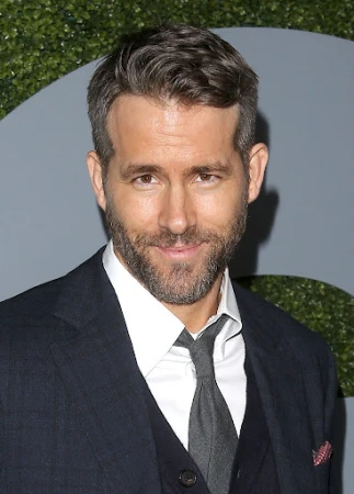 Ryan Reynolds smiling in a dark suit with a white shirt and grey tie against a patterned grey backdrop