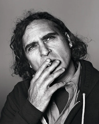 Black-and-white portrait of a middle-aged man with long hair smoking a cigarette, captured in a close-up against a neutral grey backdrop.