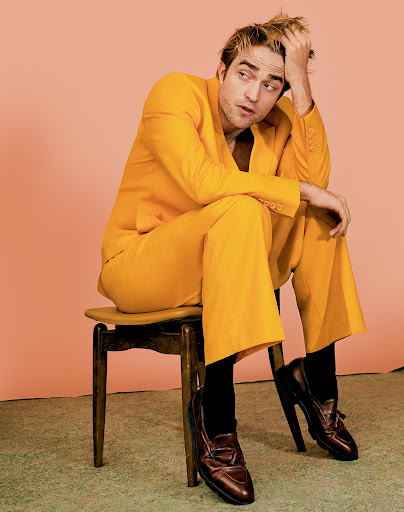 Robert Pattinson is sitting on a chair in a yellow suit with black socks and brown shoes against a peach-colored studio backdrop.