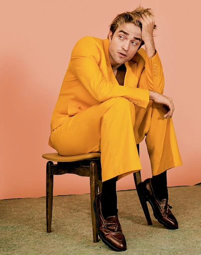 Robert Pattinson is sitting on a chair in a yellow suit with black socks and brown shoes against a peach-colored studio backdrop.