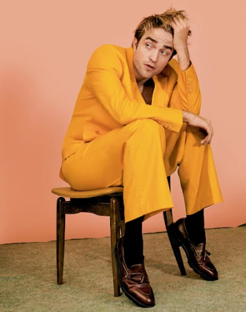 Robert Pattinson is sitting on a chair in a yellow suit with black socks and brown shoes against a peach-colored studio backdrop.