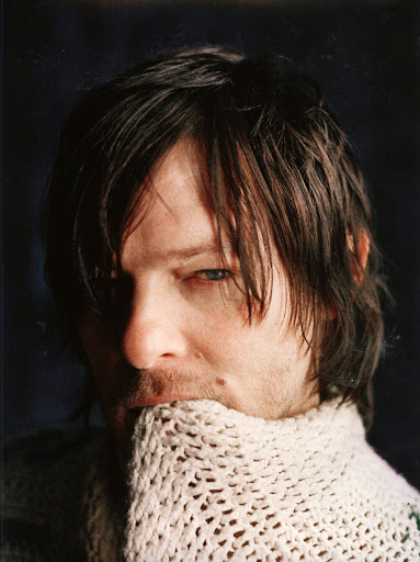 Portrait of an adult male with long dark hair and blue eyes wearing a textured, oversized knit sweater against a dark background.