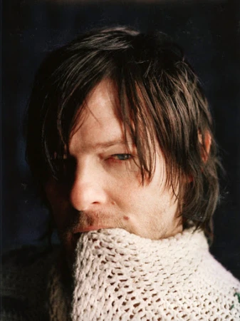 Portrait of an adult male with long dark hair and blue eyes wearing a textured, oversized knit sweater against a dark background.