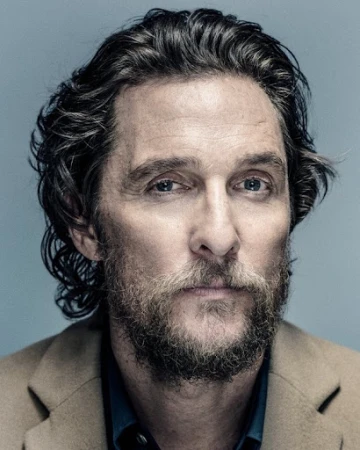 Headshot of a mature adult man with a beard and medium-length hair, wearing a collared shirt and a beige blazer against a solid backdrop.