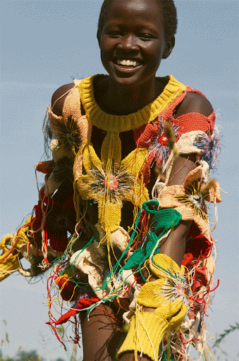 Young African woman smiles outdoors wearing a colourful textured knit dress with flower embellishments, against a blue sky and greenery.