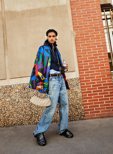 Woman in a colorful jacket, blue jeans, and black sneakers, standing outside against a textured wall, holding a purse and water bottle.