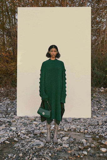 A woman stands in an outdoor fashion shot wearing a green sweater dress, carrying a matching purse, posing against a stone and white backdrop.