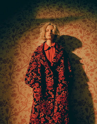 A fair-skinned woman with blonde hair in a patterned coat stands in front of floral wallpaper in a warm-toned, indoor fashion portrait.