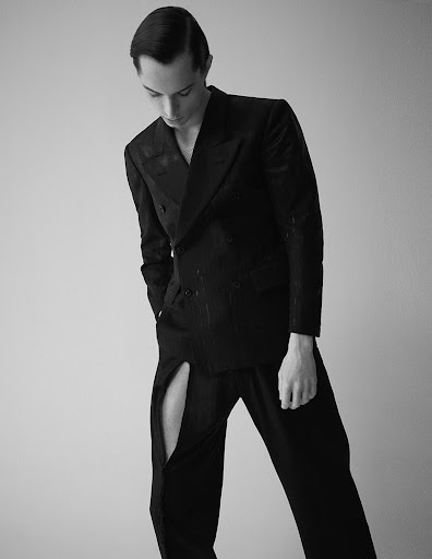 Monochrome portrait of a male fashion model in a dark suit with a slit in the pant leg, looking down against a light grey background.
