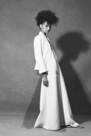 Monochrome fashion portrait of a model with voluminous hair wearing a white blazer and long skirt against a textured grey background.