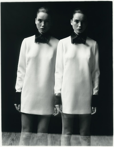 Monochrome portrait of twin women in white dresses with black collars and cuffs, mirrored on a wood surface against a dark backdrop.