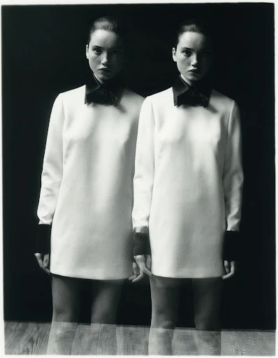 Monochrome portrait of twin women in white dresses with black collars and cuffs, mirrored on a wood surface against a dark backdrop.
