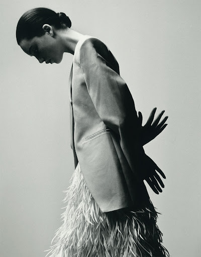 Black and white portrait of a slender woman in blazer and feathered skirt, looking down with her hands visible as shadows.