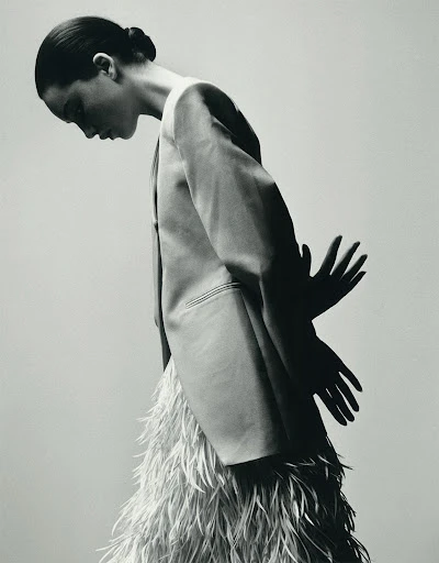 Black and white portrait of a slender woman in blazer and feathered skirt, looking down with her hands visible as shadows.