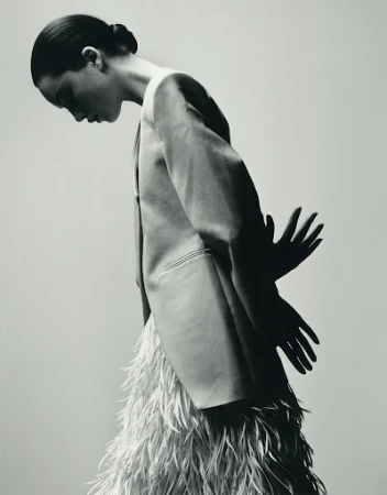 Black and white portrait of a slender woman in blazer and feathered skirt, looking down with her hands visible as shadows.