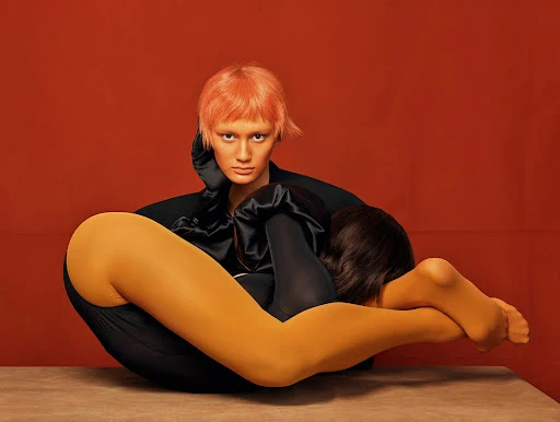 A captivating studio portrait featuring two women in a unique pose, one with peach-colored hair and the other with dark hair, against a solid orange backdrop.