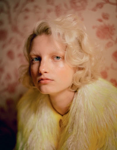 Portrait of a blonde woman with pale skin wearing a yellow fur coat, posing in an indoor setting with a floral background.