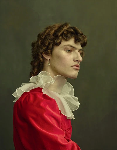 Portrait of a young adult male with curly brown hair, wearing a red top with a white ruffled collar against a dark green background.