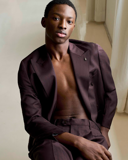 Elegant portrait of a Black male model in a dark maroon suit, posing in an indoor setting with soft, diffused natural light.