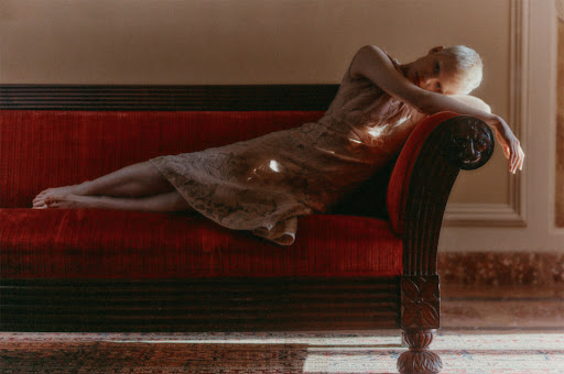 Woman with short blonde hair reclines on a red couch in dress with sunlight filtering into the room in a high-angle shot. Interior scene.