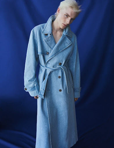 Adult male fashion model in a light-blue denim trench coat poses against a dark-blue backdrop in a studio setting with moody lighting.